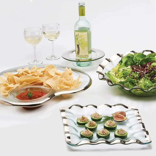 A set of elegant glass serving dishes with scalloped silver rims, styled with food on a white background. The collection includes a large oval platter with tortilla chips and salsa, a bowl of mixed green salad, a square platter with appetizers, and a small round dish holding a bottle of white wine and two wine glasses.