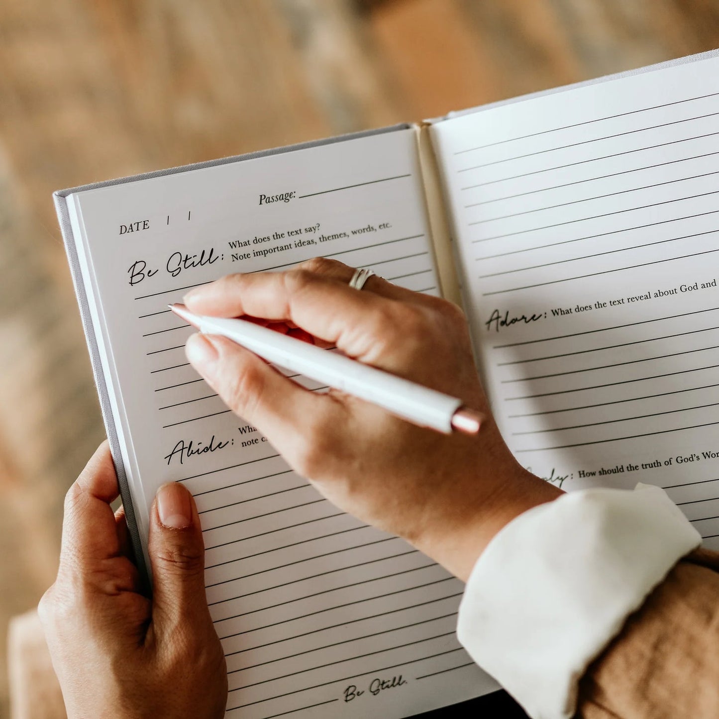 A close-up of a person's hands holding an open lined journal and writing with a white pen. The pages show handwritten notes and the title 'Be Still' at the top.