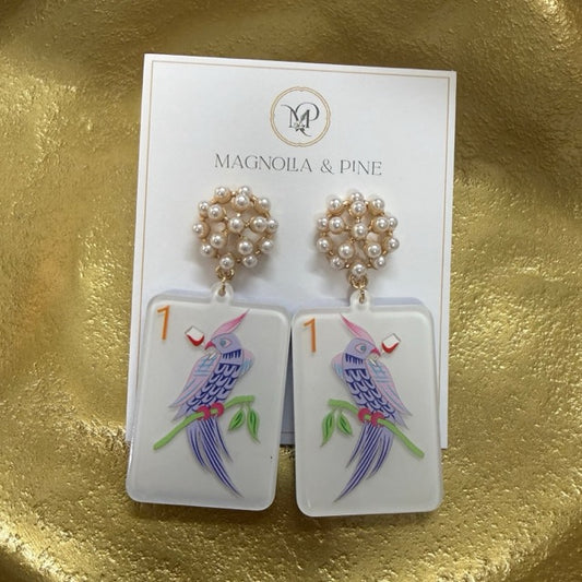 A pair of Magnolia & Pine dangle earrings displayed on their white brand card, set against a textured gold background. The earrings feature white rectangular pendants decorated with hand-painted blue birds, green leaves, and small red accents, hanging from gold-toned rings adorned with white faux pearls.