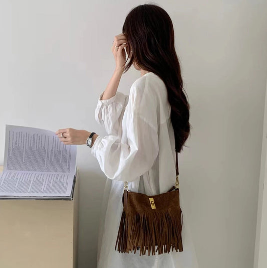 Woman in a white blouse and jeans holding a brown fringed bag, standing in front of a white wall.