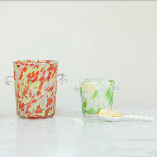 Two decorative glass ice buckets with side handles on a white marble surface against a white background. The left bucket is speckled with red, orange, and yellow on a pale green base, while the right bucket is speckled with green and yellow.