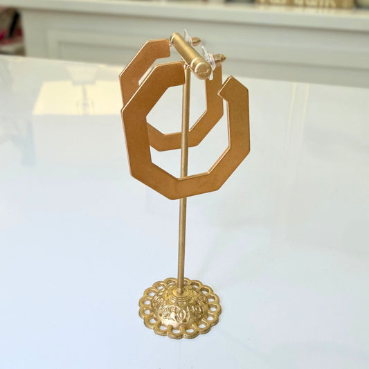 Gold-toned metal jewelry display stand with a geometric openwork top and round decorative base, photographed on a white tabletop.
