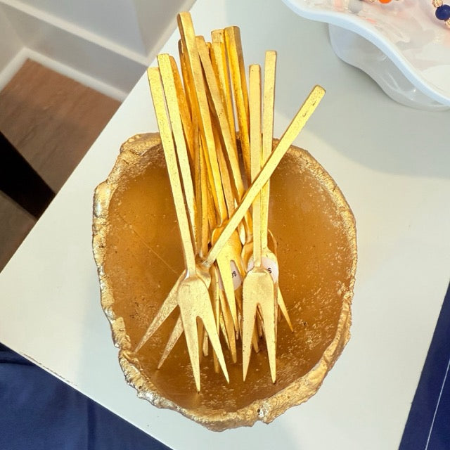 A gold-colored ceramic bowl filled with multiple gold-colored forks, photographed from a top-down angle on a white tabletop.
