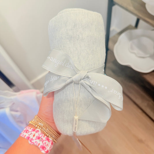 A light gray baby blanket rolled up and tied with a matching gray ribbon printed with the brand name 'angel dear', held by a hand with gold and pink bracelets.