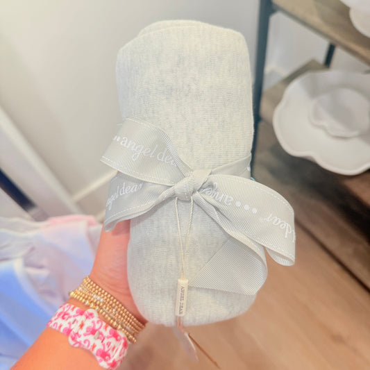 A light gray baby blanket rolled up and tied with a matching gray ribbon printed with the brand name 'angel dear', held by a hand with gold and pink bracelets.