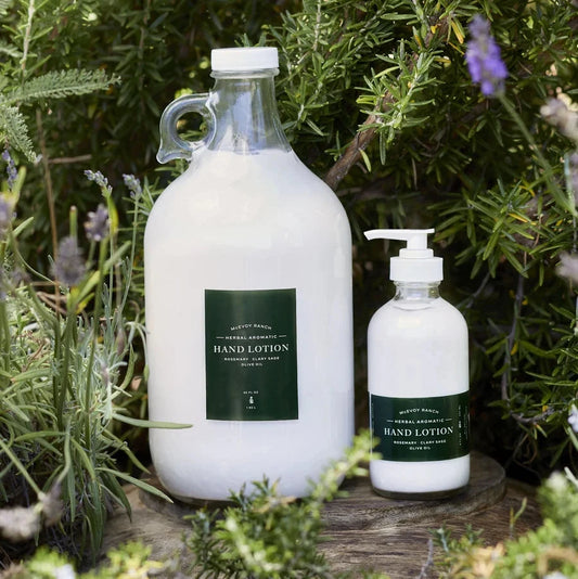 Two white hand lotion bottles from the brand 'McEvoy Ranch' displayed outdoors on a rustic wooden stump, surrounded by green rosemary and lavender plants. One bottle is a large glass jug and the other is a smaller pump bottle, both featuring dark green labels.
