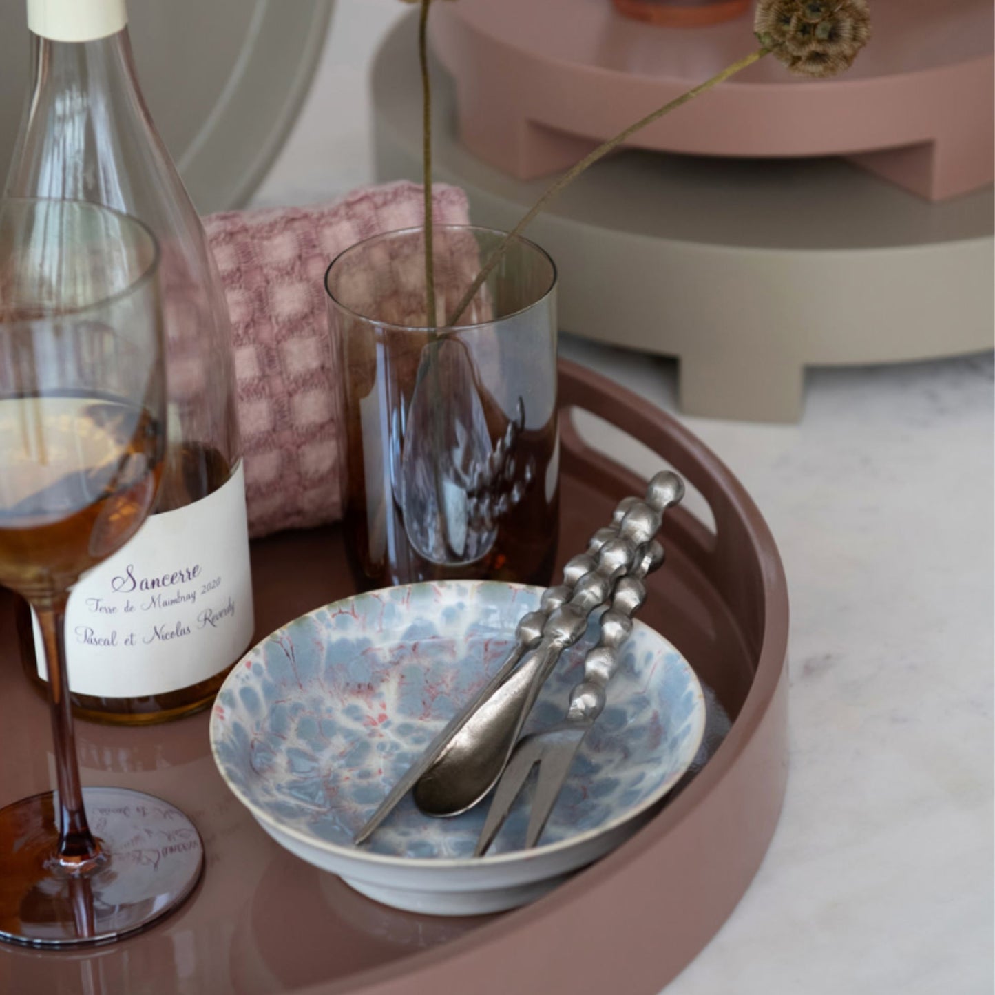 A styled tabletop scene with a round dusty-rose tray holding a blue marbled bowl, silver spoon and fork with beaded handles, a dark amber glass, and part of a wine bottle on a light marble surface.