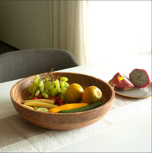 A round wooden bowl filled with green grapes, oranges, a cucumber, and small vegetables, placed on a table runner in a bright indoor setting. A plate with sliced fruit sits in the background.