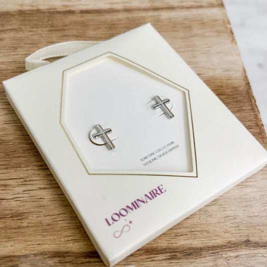 Silver cross stud earrings displayed on a cream-colored hexagonal jewelry card, which is placed on a wooden surface. The brand name 'LOOMINAIRE' is printed in purple on the card.