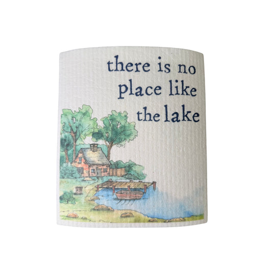 A cream Swedish dishcloth illustrated with a lakeside cottage and dock, reading 'There is no place like the lake.'