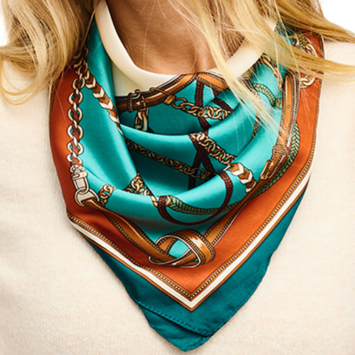 A teal silk scarf with a cognac and tan chain-link border print, knotted at the neck.