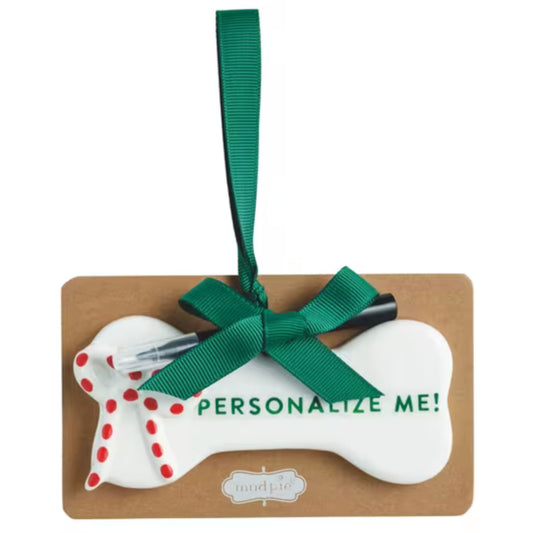 A white ceramic dog bone ornament with a red polka-dot bow design and 'Personalize Me!' text, tied with a green ribbon.