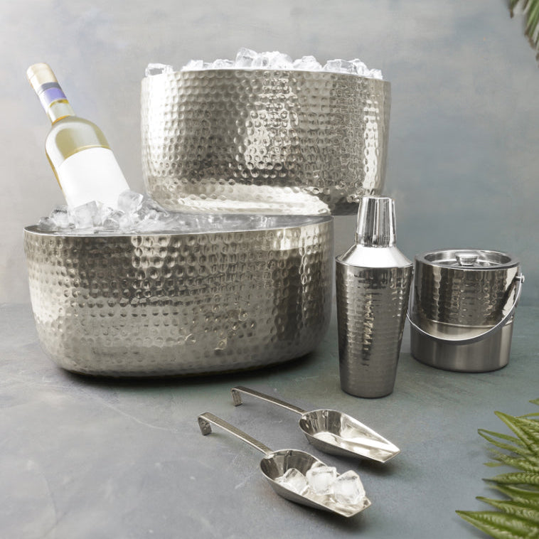 Metallic ice buckets and shakers with ice on a gray surface, surrounded by greenery.