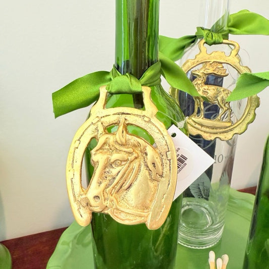 A green glass bottle decorated with a gold horseshoe-shaped horse emblem hanging from a green satin ribbon, with a white product tag attached.
