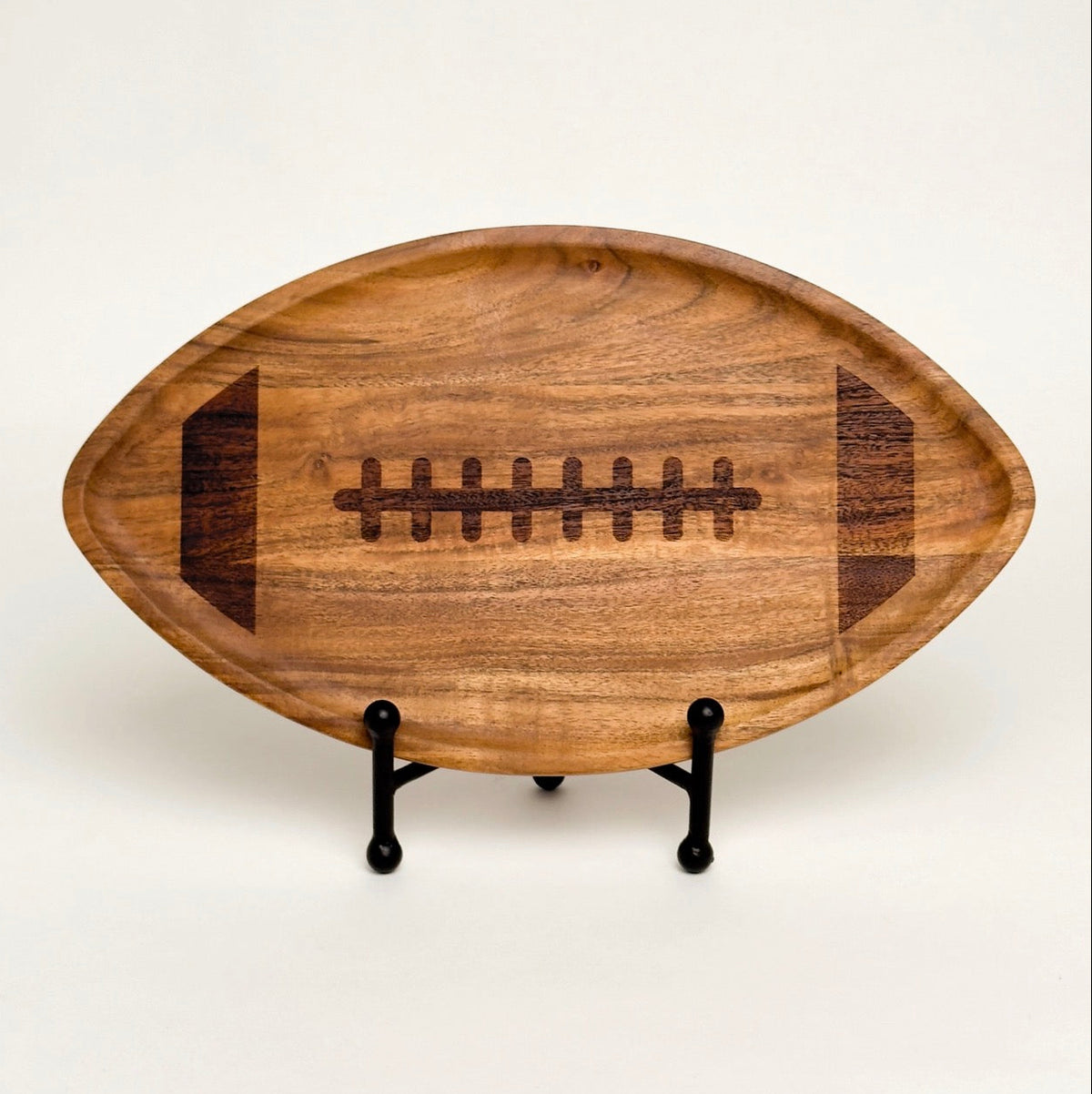 A wooden football-shaped serving tray with a dark engraved laces design, displayed on a small black metal stand against a plain off-white background.