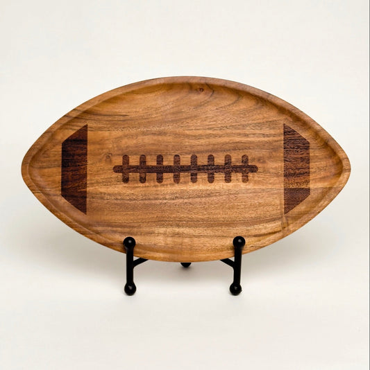 A wooden football-shaped serving tray with a dark engraved laces design, displayed on a small black metal stand against a plain off-white background.