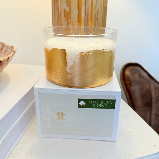 Candle with gold base and white wax on a box labeled 'Magnolia & Pine' on a decorative surface.