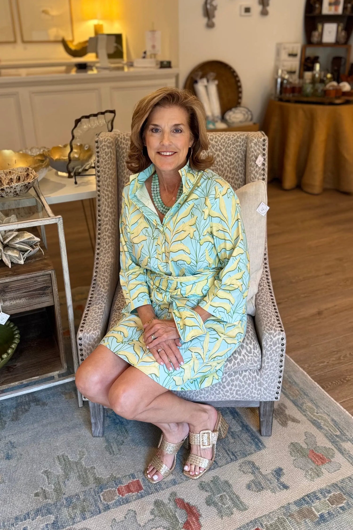 Picture of Magnolia & Pine owner, Hope Holt, wearing a light blue and yellow tropical print dress sits in a gray upholstered armchair inside a store setting with wood floors and home decor.
