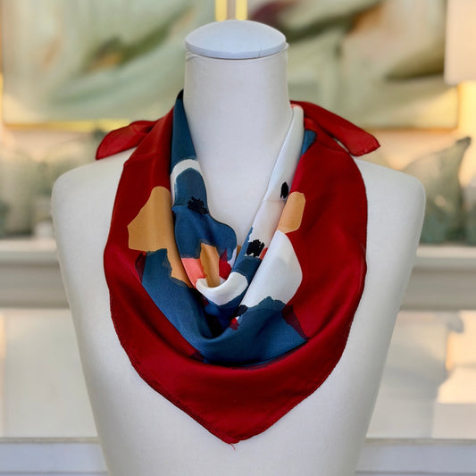 A multicolor silk scarf with a red base and abstract blue, white, and yellow patterns, styled on a white mannequin torso in a brightly lit room.