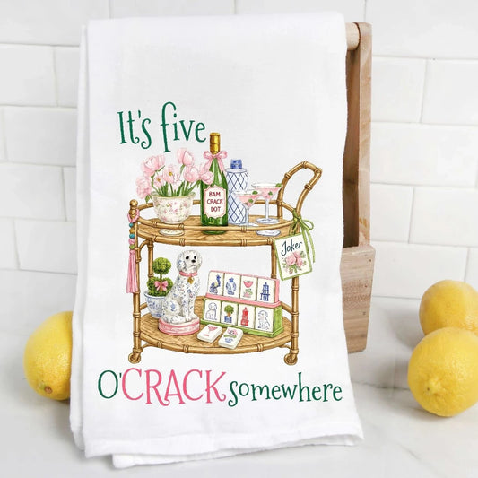 A white kitchen towel with a colorful cartoon illustration of a bar cart and the text 'It's five O'CRACK somewhere' is draped over a wooden stand. The towel is styled on a white marble countertop with yellow lemons on both sides against a white subway tile backsplash.