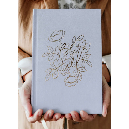 A person holds a light blue hardcover journal with gold foil floral line art and the words 'Be Still' on the front cover.
