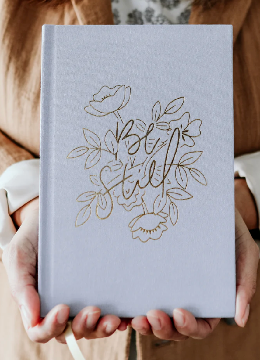 Person holding a light blue notebook with 'Be Still' and floral designs.