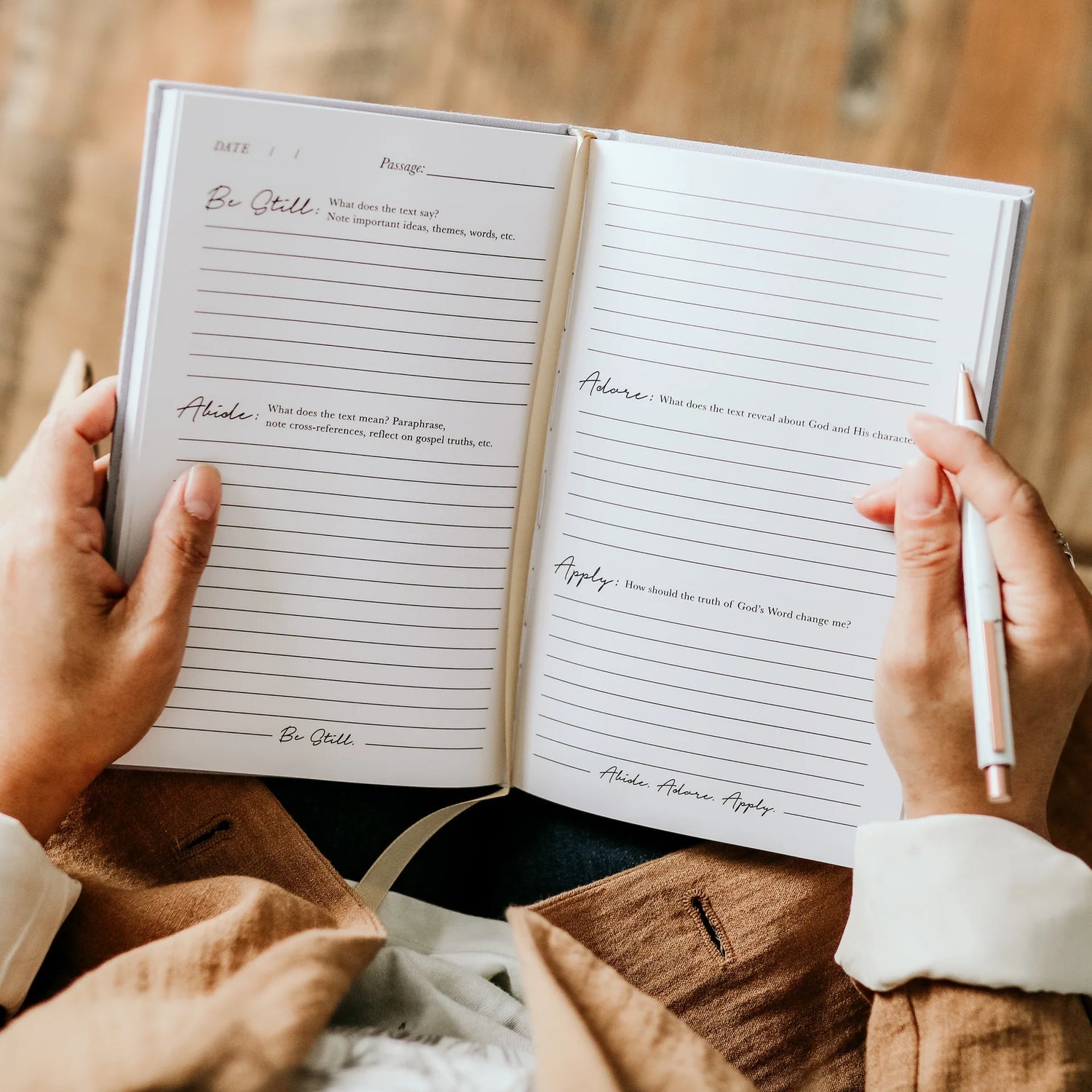 An open lined journal with handwritten notes and a white pen held in both hands, set against a warm, blurred background of clothing and wood.