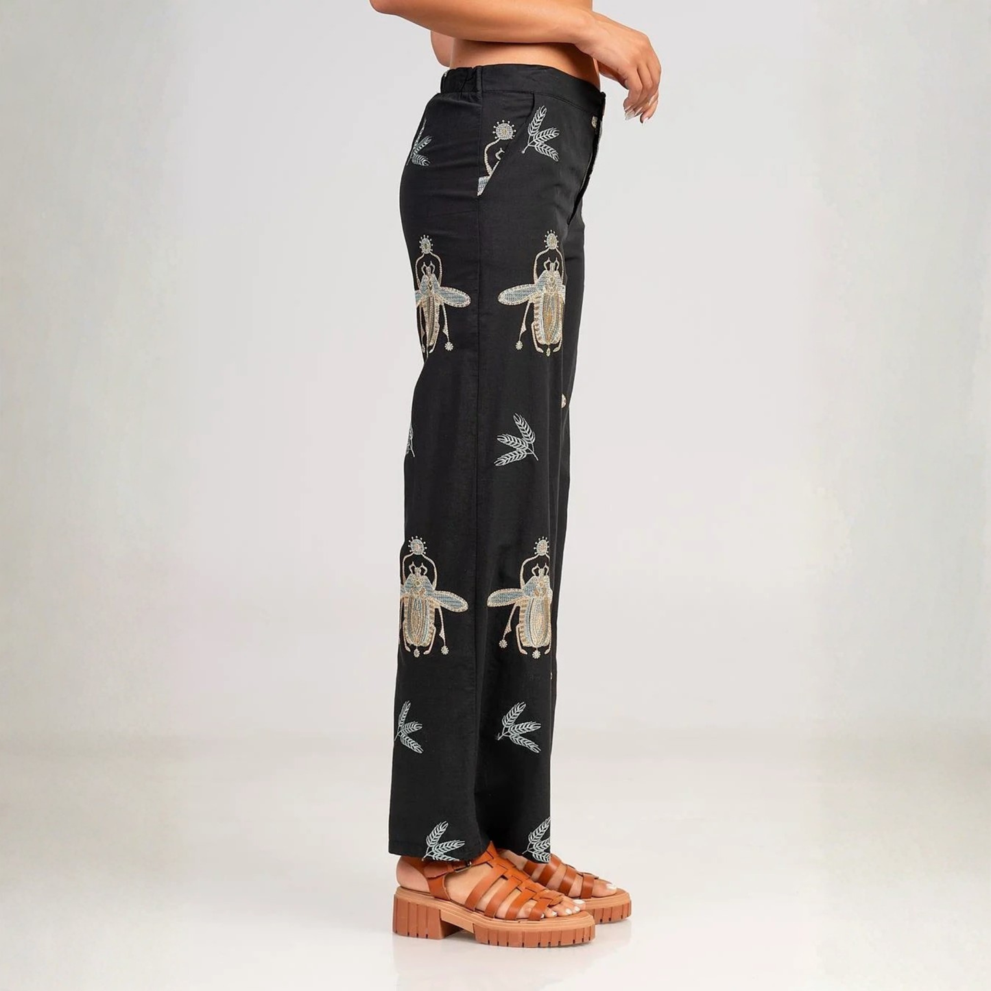 A model is shown from the waist down wearing black wide-leg trousers with embroidered beetles, paired with brown leather slide sandals, against a light gray studio background.