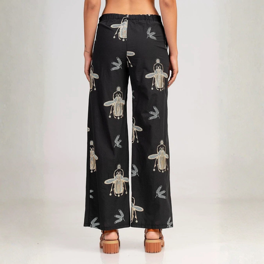 Black wide-leg pants with a beetle pattern, shown from the back on a model wearing brown platform sandals against a light gray studio background.