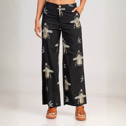 A model wears wide-leg black pants featuring a repeating pattern of embroidered-style beetle, paired with brown platform sandals against a light gray studio background.