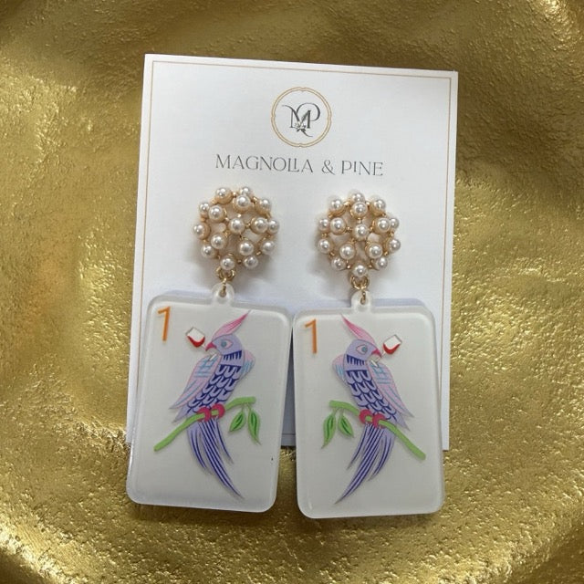 A pair of Magnolia & Pine dangle earrings displayed on their white brand card, set against a textured gold background. The earrings feature white rectangular pendants decorated with hand-painted blue birds, green leaves, and small red accents, hanging from gold-toned rings adorned with white faux pearls.