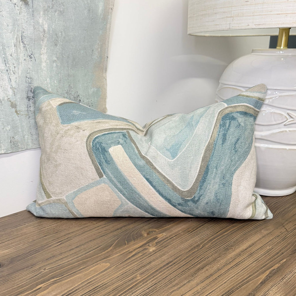 A decorative lumbar throw pillow with an abstract marbled pattern in shades of blue, beige, and gray, styled on a wooden floor next to a white ceramic lamp.