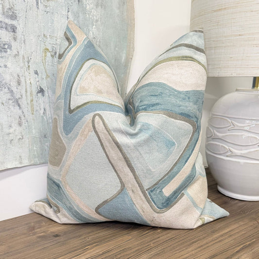 A square decorative throw pillow with an abstract geometric pattern in shades of blue, beige, and gray, styled on a wooden surface next to a white ceramic lamp.