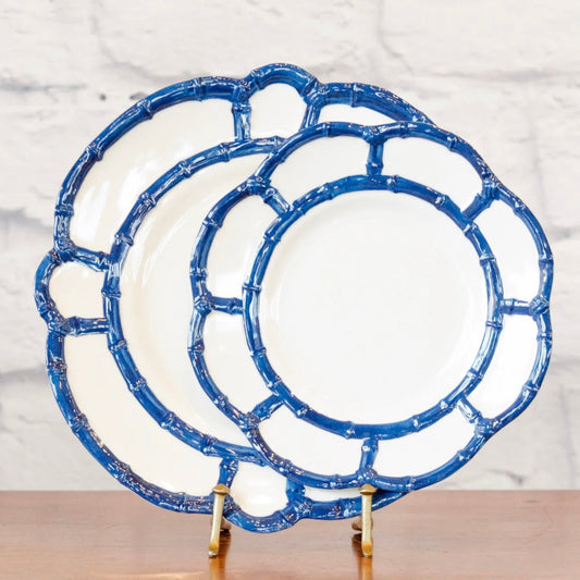 Three round ceramic plates with scalloped edges and a blue bamboo-inspired rim pattern, displayed on small gold stands against a white brick wall background.
