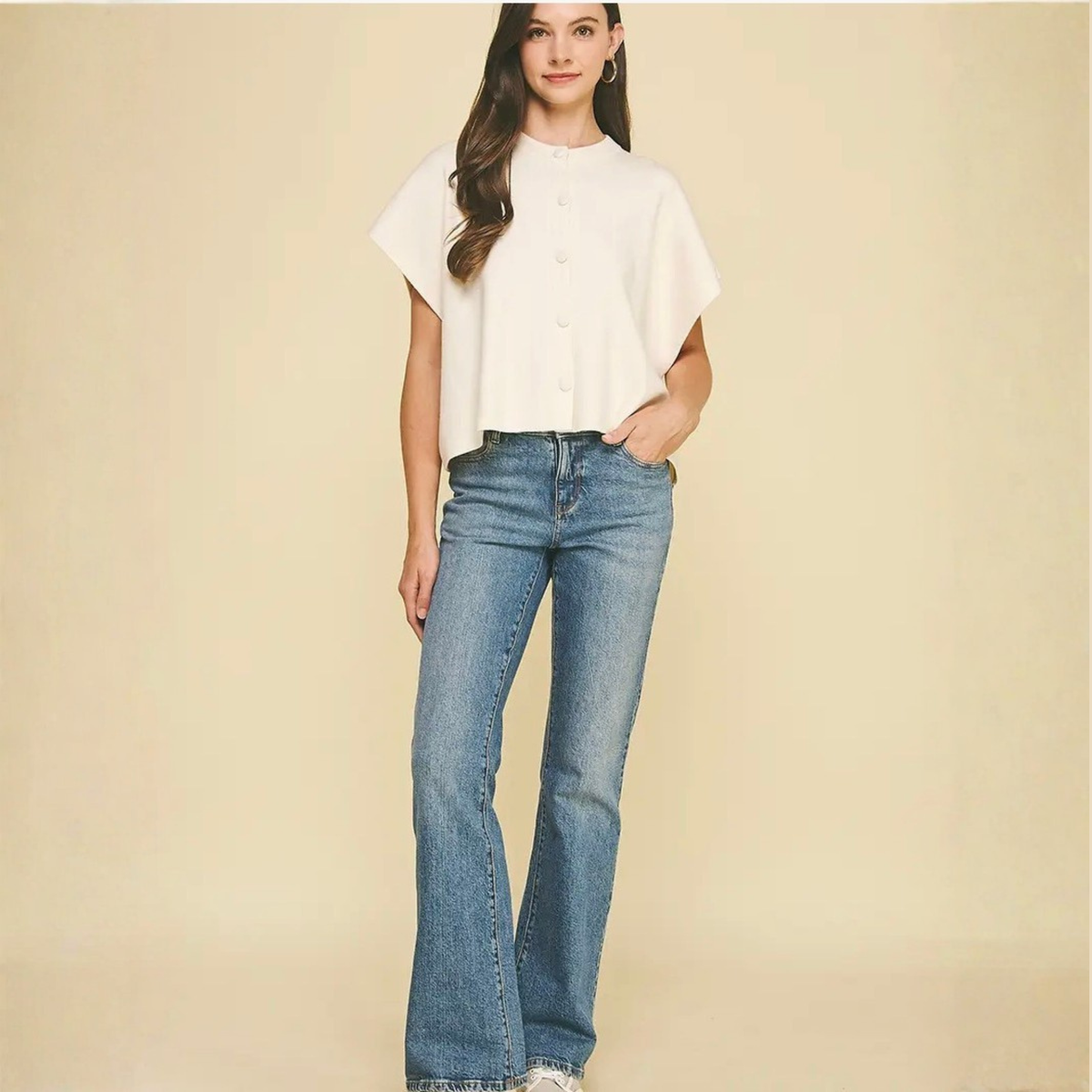 A woman modeling a white short-sleeve button-front blouse with a relaxed fit and blue flare jeans, standing against a plain beige studio background.