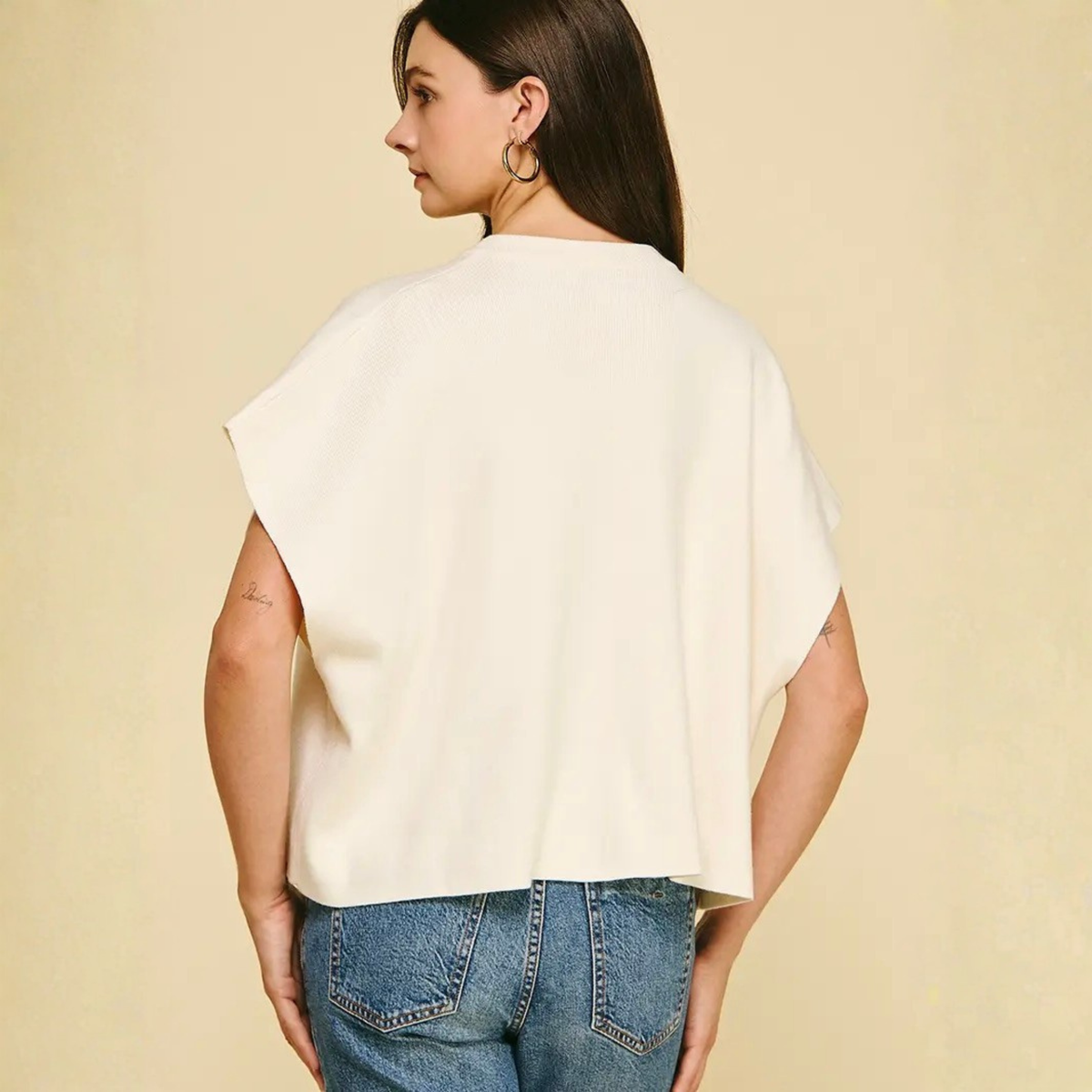 A woman shown from the back wearing a cream-colored, short-sleeved draped top and blue jeans against a solid beige background.
