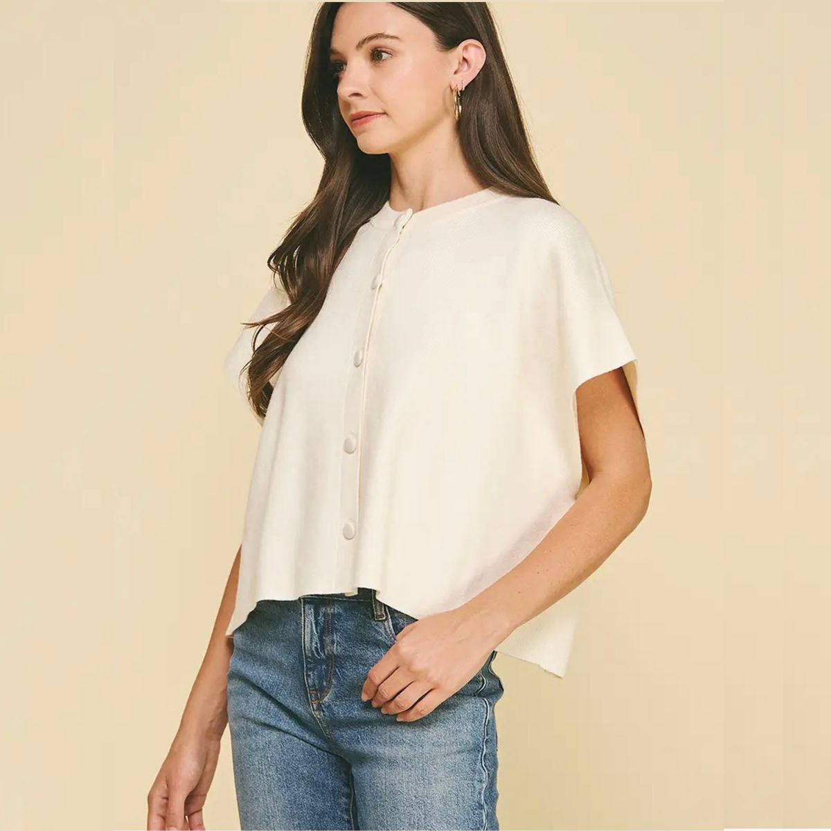 A woman wearing a short-sleeve cream-colored blouse with a button-up front and blue jeans, standing against a solid light beige background.