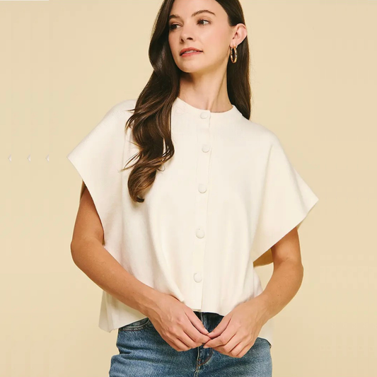 A woman wearing a cream-colored short-sleeve button-up blouse with a high mock neck, paired with blue jeans, standing against a solid beige background.