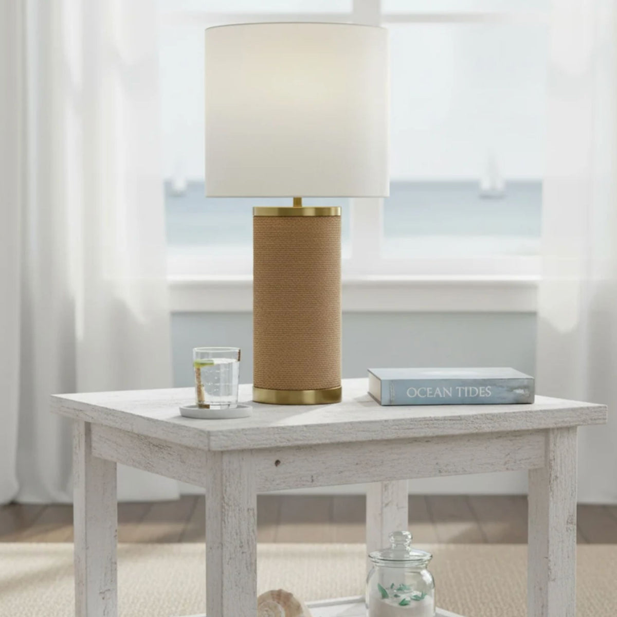 A table lamp with a natural abaca rope-wrapped base, gold accents, and a white drum shade, styled on a side table near a window.
