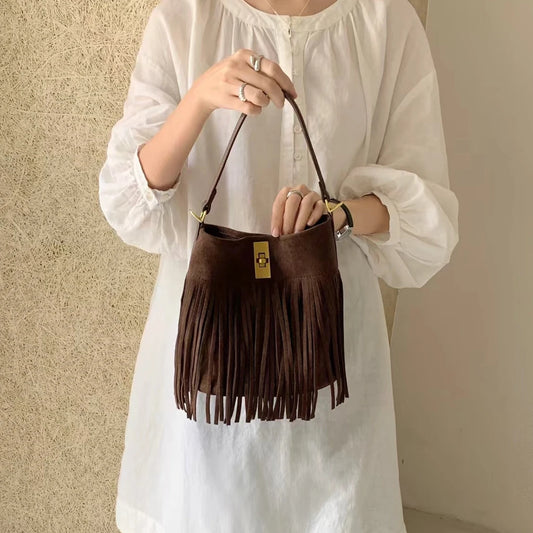 A person wearing a white long-sleeve blouse and light blue jeans holds a brown fringe shoulder bag with a gold-tone clasp against a textured beige wall.