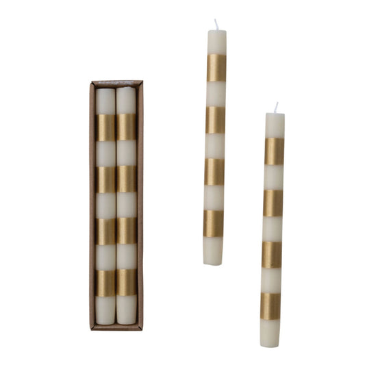 A pair of cream and metallic-gold striped taper candles standing upright, with two more candles laid beside a slim brown box.