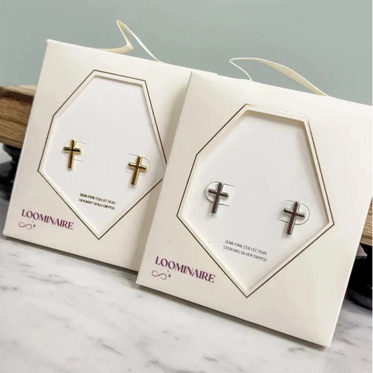 Two white LOOMINAIRE jewelry display cards, each holding a small cross stud earring, placed on a light marble surface against a pale gray wall.