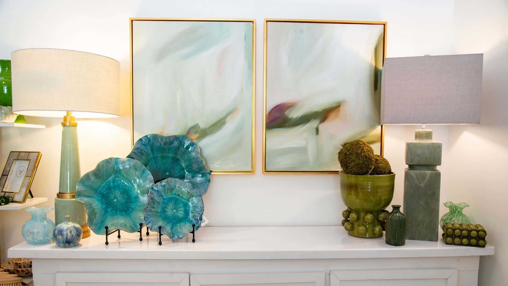 "A styled interior of Magnolia & Pine showing two framed abstract wall art prints hanging above a white console table. The artwork has soft white, pale blue, and gray tones with subtle green and purple accents."