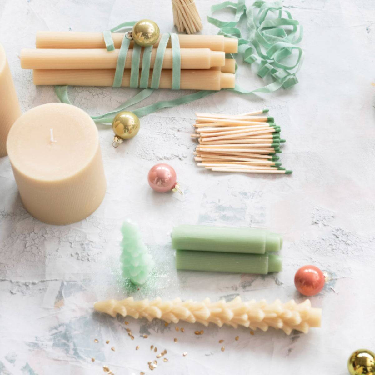 A cream tree-shaped taper candle lying on a holiday flat lay with a pillar candle, bundled cream tapers tied in green ribbon, sage green tapers, matchsticks, and pink and gold ornaments.
