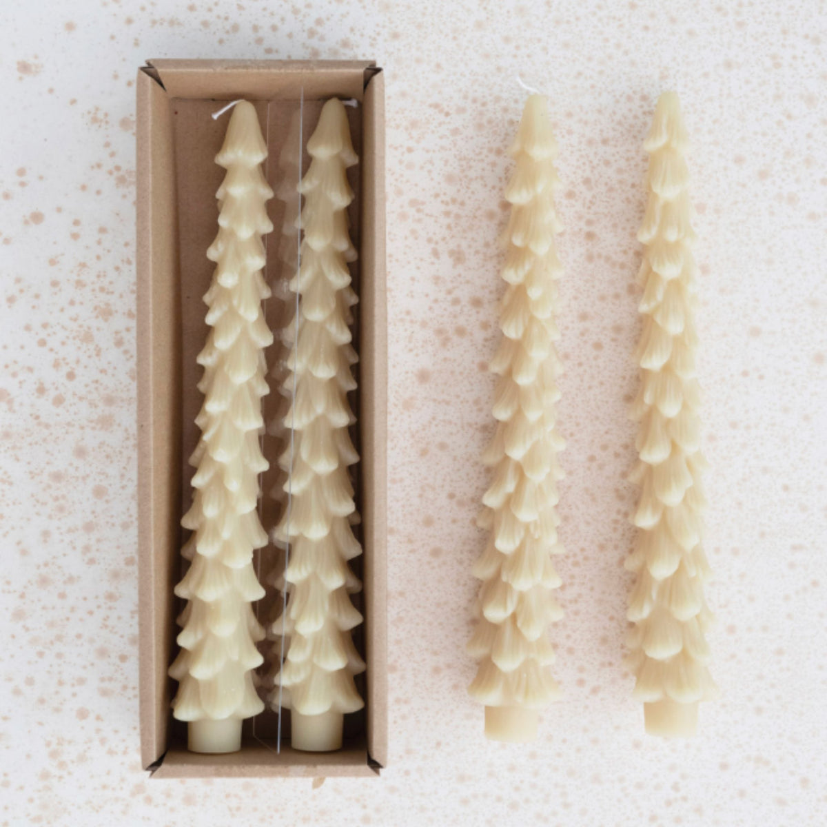 A kraft box holding two cream tree-shaped taper candles, shown next to two more of the candles out of the box on a speckled white surface.