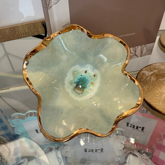 A ceramic flower-shaped dish with a wavy gold rim and a pale green to blue-green glazed interior, displayed on a glass shelf with other decorative items around it.