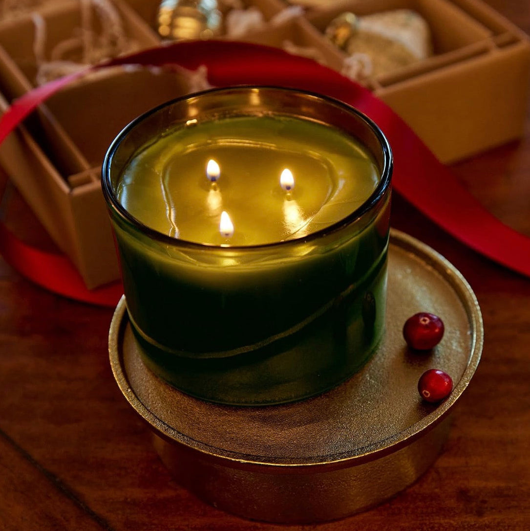 A lit three-wick candle in a dark green glass jar sits on a round metal tray on a wooden table, with a kraft gift box, red ribbon, and small red berries in the background.