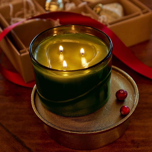 A lit three-wick candle in a dark green glass jar sits on a round metal tray on a wooden table, with a kraft gift box, red ribbon, and small red berries in the background.