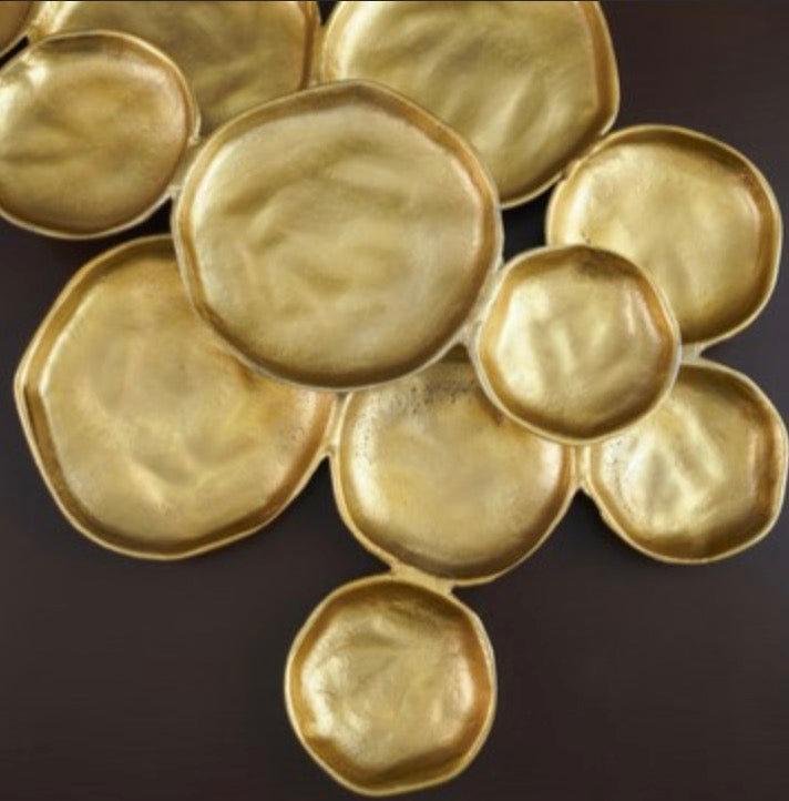 A cluster of organic-shaped gold metallic dishes or trays with a textured, hammered finish, arranged on a dark brown background.