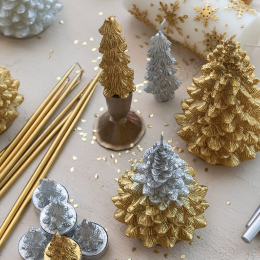 A styled flat-lay of metallic Christmas tree ornaments in gold and silver, including several miniature pine tree candle holders, a few gold taper candles, and a white candle with gold glitter snowflakes on a neutral beige background.